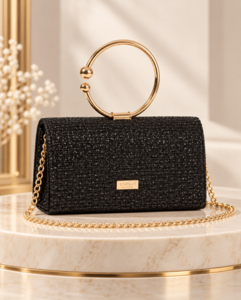 Clutch Morgan Tijou negro de acabado brillante y forma rectangular, con asa circular dorada, cadena metálica y placa decorativa frontal, sobre una mesa de mármol claro.