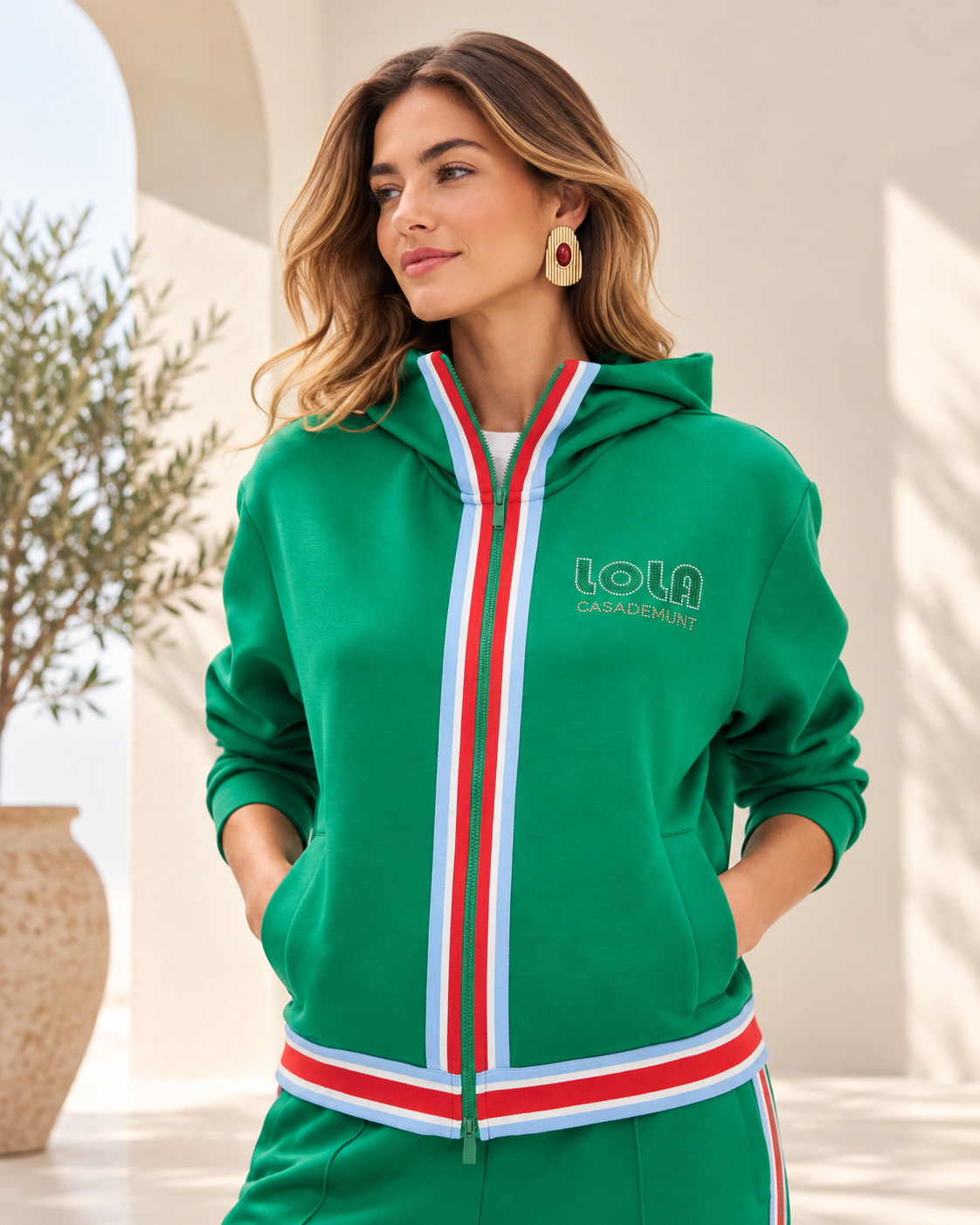 Retrato editorial en chándal verde mediterráneo Modelo con sudadera Lola Casademunt verde de capucha y cremallera, detalles tricolor en blanco y rojo, logotipo bordado y bolsillos delanteros.